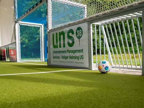 Ein bunter Ball liegt neben einem blauen Fußballtor sowie einem Schild mit der Aufschrift "uns 38" auf Kunstrasen.