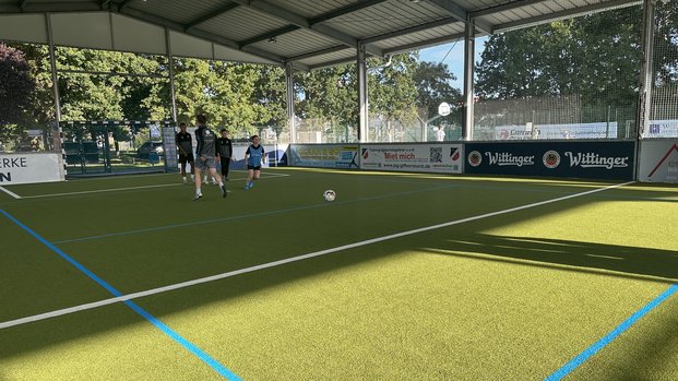 Personen spielen Fußball in einer überdachten Halle mit Kunstrasen und blauen Linien.