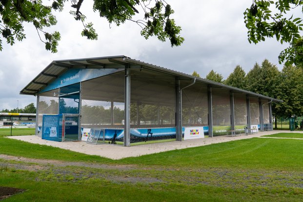"Moderne Sportarena im Grünen: Die BraWo McArena im Freien" Überdachtes Sportkäfigfeld mit Werbebannern, umgeben von grünem Rasen und Bäumen.