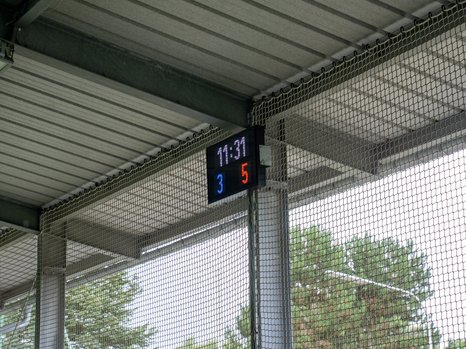 Ein digitaler Anzeigetafel in einem überdachten Bereich zeigt die Uhrzeit 11:31 und den Spielstand 3 zu 5. Die Tafel ist an einem Metallrahmen montiert, und der Bereich ist durch Maschendraht gesichert. Im Hintergrund sind Bäume sichtbar, die einen Teil der Außenlandschaft zeigen.