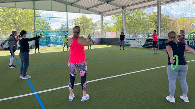 "Gemeinschaftliches Workout in der BraWo McArena" Menschen machen Übungen mit grünen Ringen in einer überdachten Sportanlage bei Tageslicht.