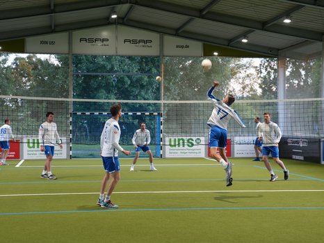 Eine Gruppe von Männern spielt Faustball in einer Halle. Einer der Spieler springt in die Luft und bereitet sich darauf vor, den Ball mit der ausgestreckten rechten Hand zu schlagen. Die anderen Spieler beobachten das Geschehen, während sie sich auf dem Spielfeld positionieren. Die Halle hat eine hohe Decke und auf den Wänden sind Werbebanner sichtbar.