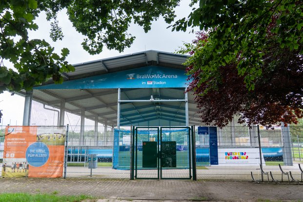 "BraWo McArena: Ein Ort für Sport und Gemeinschaft" Überdachte Sporthalle mit Netz, Bäumen im Vordergrund und Schildern „BraWo McArena“ und „Die Halle für Alle(s)“.
