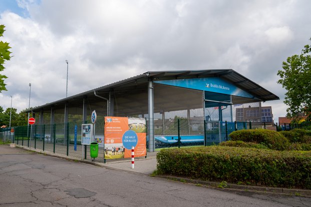 "Moderne Sporthalle: Die BraWo McArena im Freien" Eine überdachte Sporthalle mit dem Namen "BraWo McArena" ist von einem Zaun umgeben. Ein Werbebanner an der Vorderseite wirbt für die Halle mit der Aufschrift "Die Halle für alle!" Es stehen Informations- und Anmeldehinweise auf dem Banner. Vor der Halle befindet sich eine gepflasterte Fläche mit einem Mülleimer und einem Verbotsschild für die Einfahrt. Im Hintergrund sind einige Bäume sowie ein Gebäude mit Solarpanelen auf dem Dach zu sehen. Wolken bedecken den Himmel.