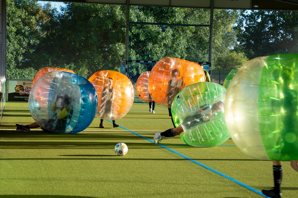 "Bubble-Fußball: Spaß und Action auf dem Spielfeld" Spieler in aufblasbaren Kugeln spielen auf einem Spielfeld mit einem Fußball, umgeben von Bäumen und einem Netz.