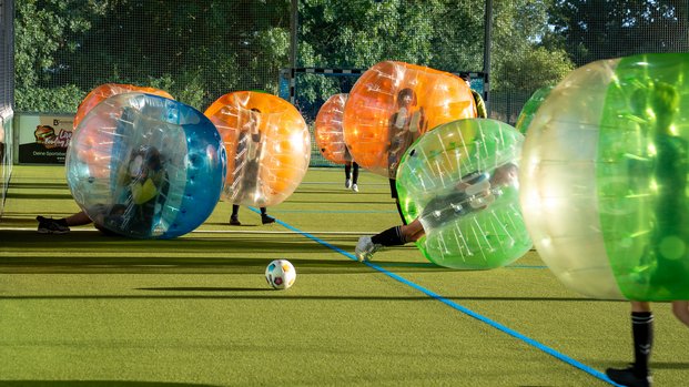 "Bubble-Fußball: Spaß und Action auf dem Spielfeld" Spieler in aufblasbaren Kugeln spielen auf einem Spielfeld mit einem Fußball, umgeben von Bäumen und einem Netz.