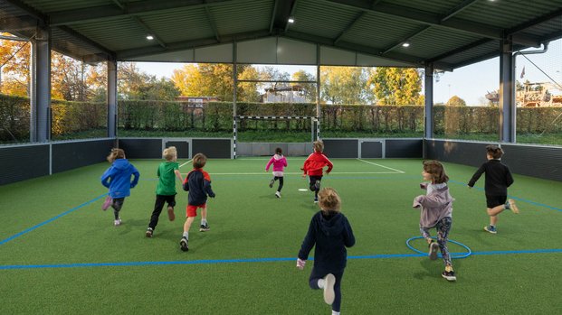 Kinder spielen in einer überdachten, umzäunten Sportanlage auf einem grünen Kunstrasenplatz.