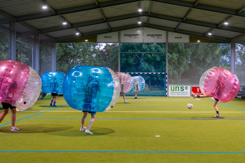 "Bubble Soccer: Spaß und Action auf dem Spielfeld" Personen in aufblasbaren Bällen spielen Bubble-Fußball in einer Halle auf Kunstrasen.